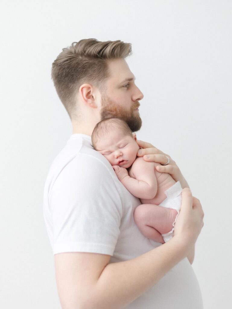 Dad in white t-shirt holds tiny newborn baby curled up on his chest.